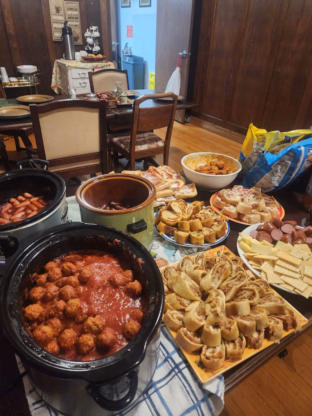 A dining area with a table set with various foods including meatballs in sauce in a slow cooker, mini sausages in another slow cooker, assorted rolled sandwiches, crackers with sliced sausage and cheese, and bags of chips. The background shows wooden paneled walls, chairs, and a small table with a coffee dispenser and cups.