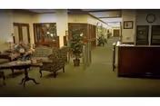 Interior view of a senior living facility hallway with a seating area on the left featuring floral arrangements and upholstered chairs, a wooden reception desk on the right, and a corridor extending into the background with plants and framed pictures on the walls.