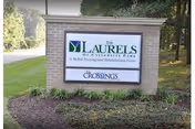 A brick sign for The Laurels of University Park, a senior living and rehabilitation center, situated outdoors with grass and trees in the background.