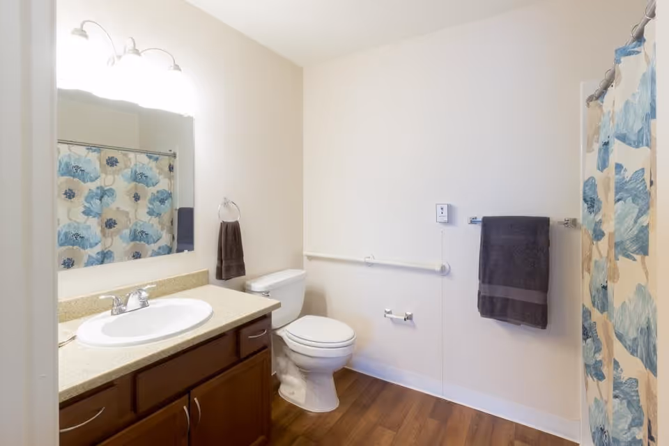 A clean bathroom with a white toilet, a sink with a beige countertop and wooden cabinets, a large mirror above the sink, a towel ring with a dark towel, a grab bar on the wall next to the toilet, a toilet paper holder, and a shower with a blue and beige floral shower curtain.
