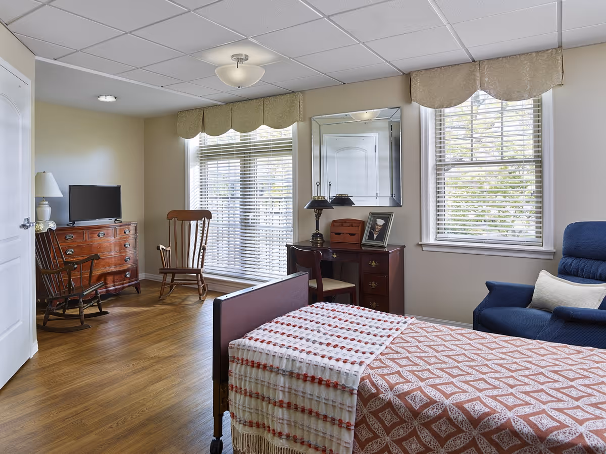 A well-lit bedroom in a senior living facility featuring a single bed with a patterned bedspread and a throw blanket. The room includes a blue armchair with a white pillow, a wooden desk with a lamp and a framed photo, a wooden rocking chair, a dresser with a TV on top, and two large windows with blinds and valances. The floor is wooden, and the walls are painted light beige.