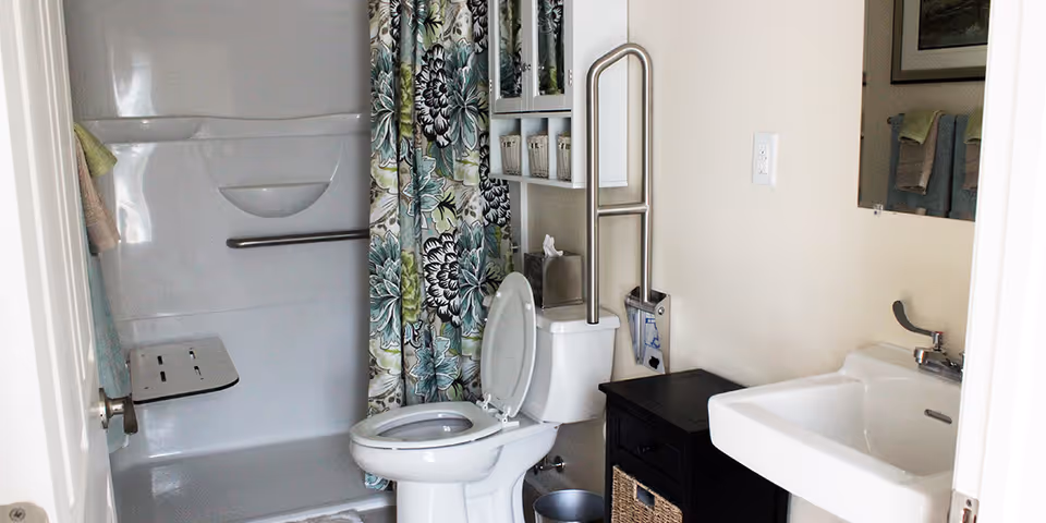 Small accessible bathroom with a toilet, sink, and a shower with a floral curtain and fold-down seat, plus grab bars and storage shelving.