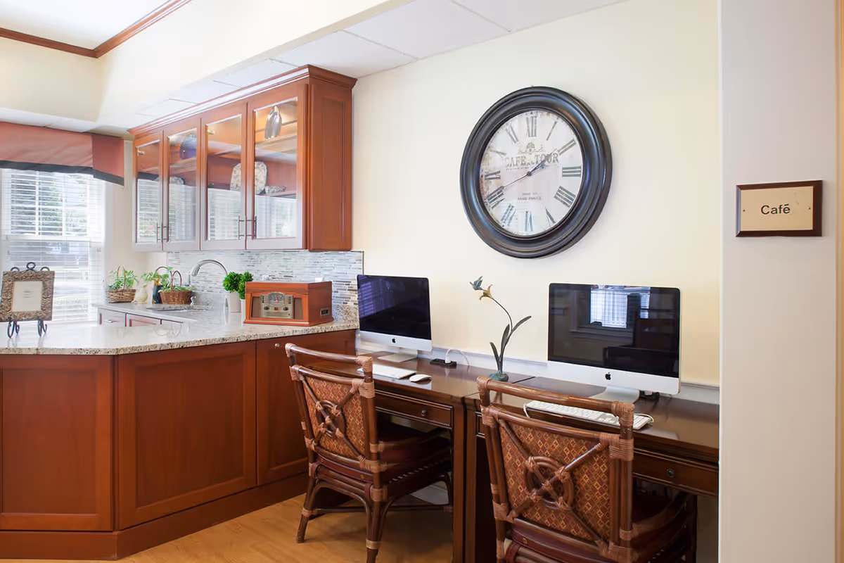 Interior view of a café area with two wooden chairs and a wooden desk holding two desktop computers. Behind the desk is a large round wall clock with Roman numerals. To the left, there is a countertop with wooden cabinets above and below, decorated with plants and a vintage radio. A window with blinds and a valance lets in natural light.