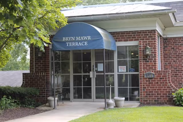 Entrance to a brick building with a blue awning that reads 'Bryn Mawr Terrace'. The entrance has glass double doors and windows on either side. There are two small planters and a bench near the door, with green grass and trees surrounding the walkway leading to the entrance.