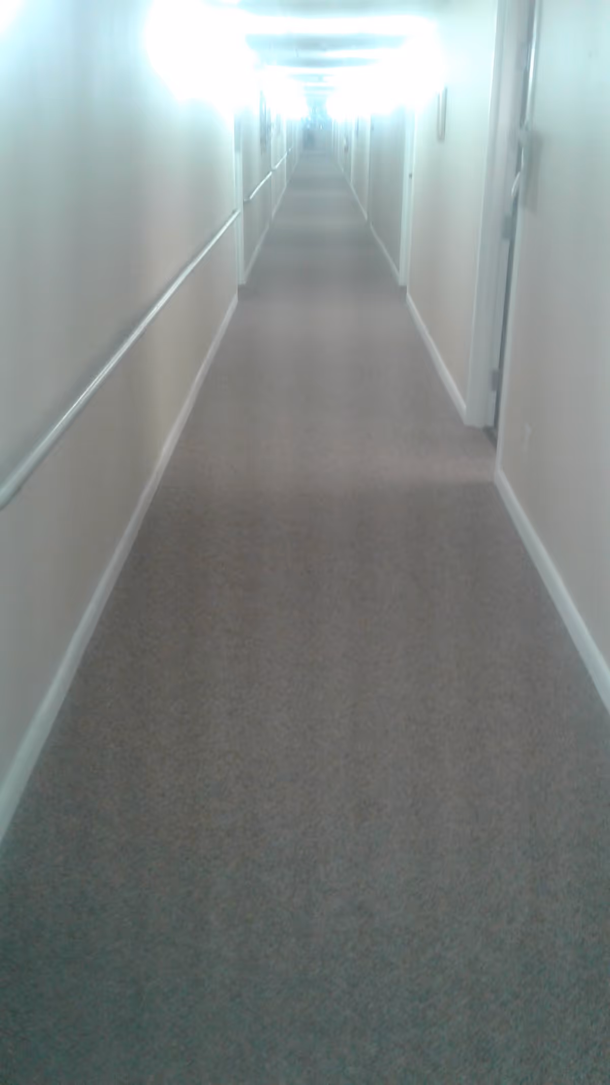 A long, narrow hallway with beige walls and carpeted floor, featuring handrails on both sides and several closed doors along the corridor, illuminated by bright overhead lights.