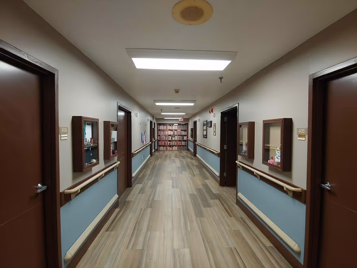 Long interior hallway with wood-look flooring, handrails, numbered resident doors and display cases, and a bookshelf at the far end.