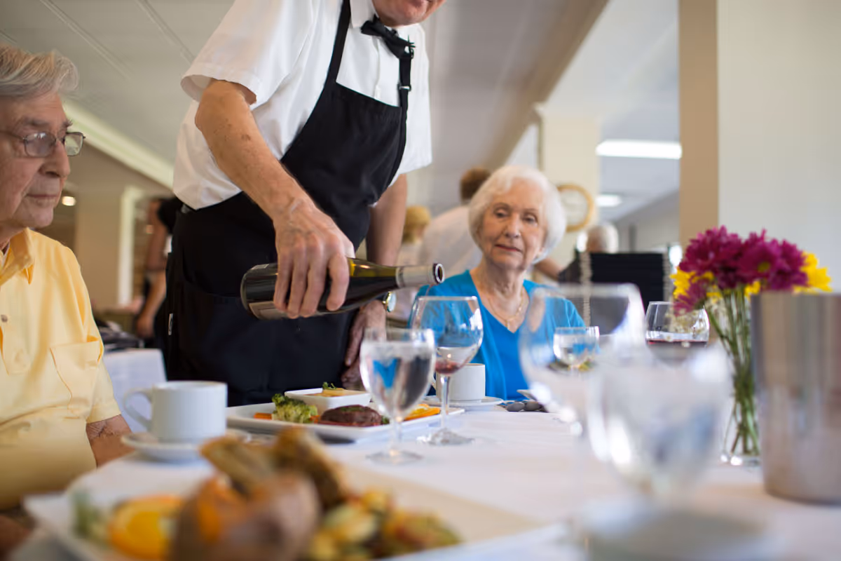A server pours wine for elderly residents seated at a dining table in a communal dining room.