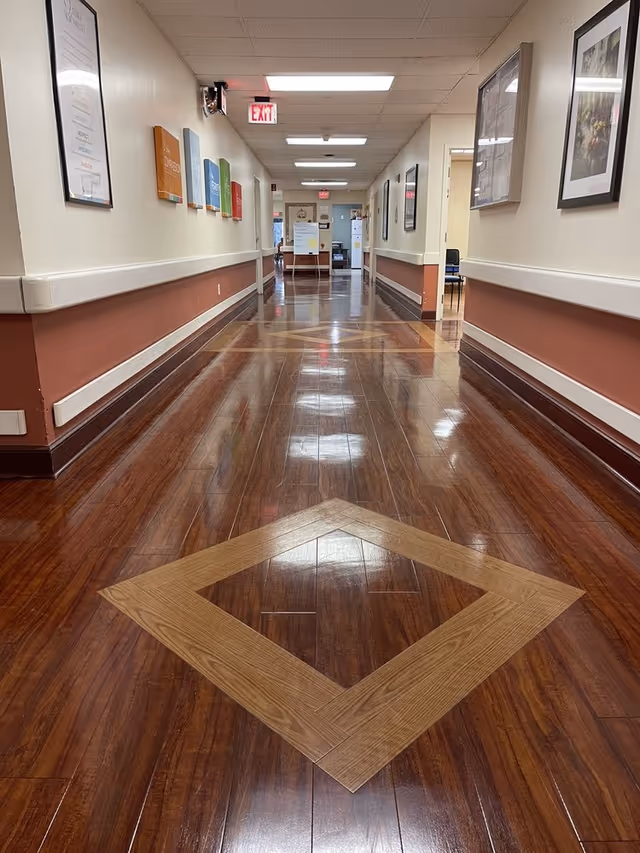 A long polished hallway inside a senior living facility with wood-patterned floors, framed artwork, handrails and an exit sign at the far end.