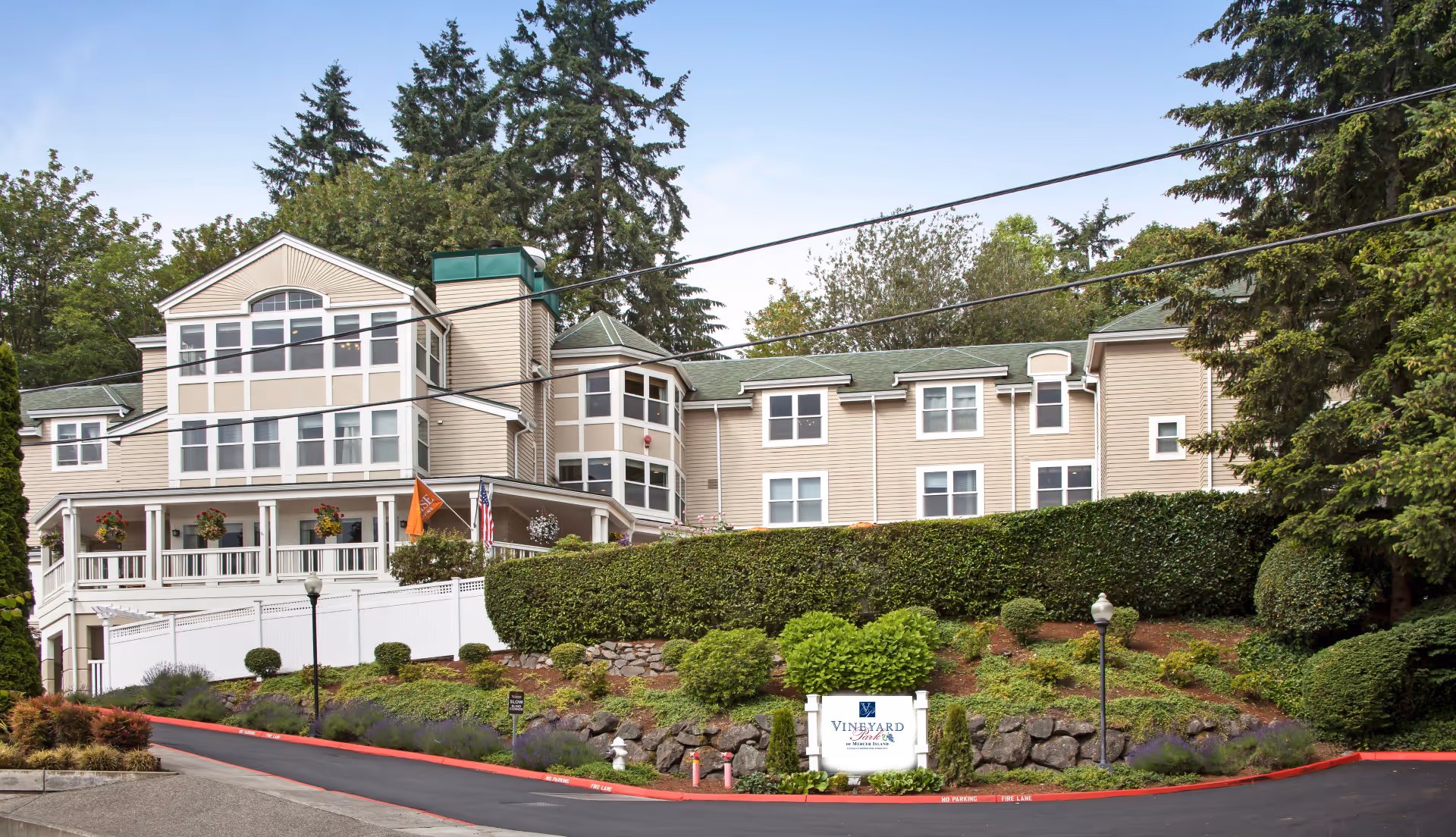 Exterior view of Vineyard Park of Mercer Island, a multi-story senior living facility surrounded by trees and landscaped greenery, with a driveway and a sign in front.