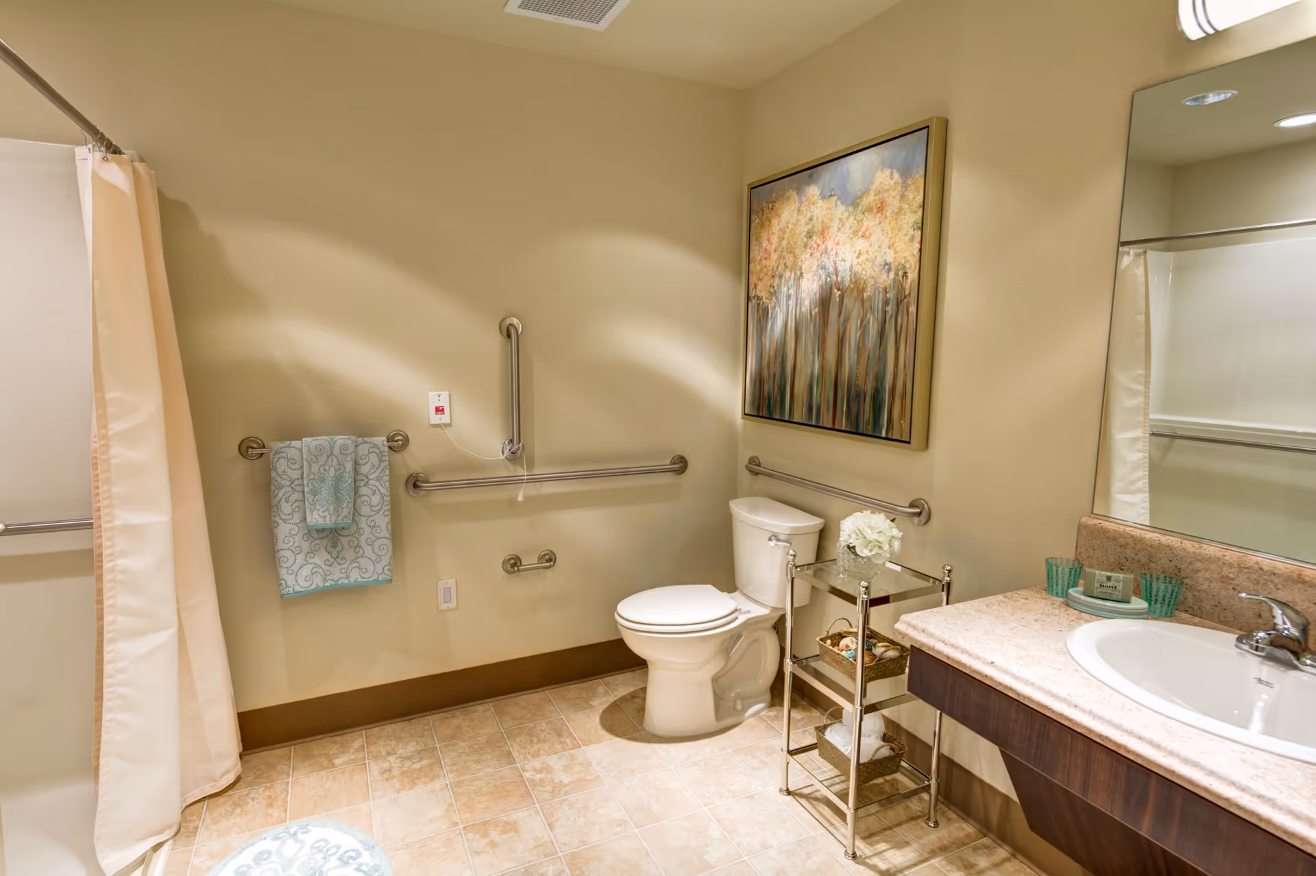 A clean and accessible bathroom with beige walls and tiled floor. The bathroom features a toilet with grab bars on the walls around it, a shower with a beige curtain, a sink with a countertop, and a small metal shelf holding decorative items and towels. A framed painting of trees hangs on the wall above the toilet.