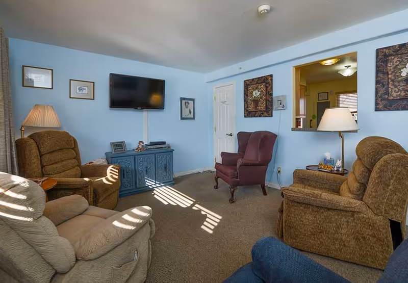 A cozy living room with light blue walls, featuring four armchairs arranged around a blue cabinet with a flat-screen TV mounted above it. The room has framed pictures on the walls, a floor lamp, and a pass-through window to an adjacent room with yellow walls and a window with blinds.