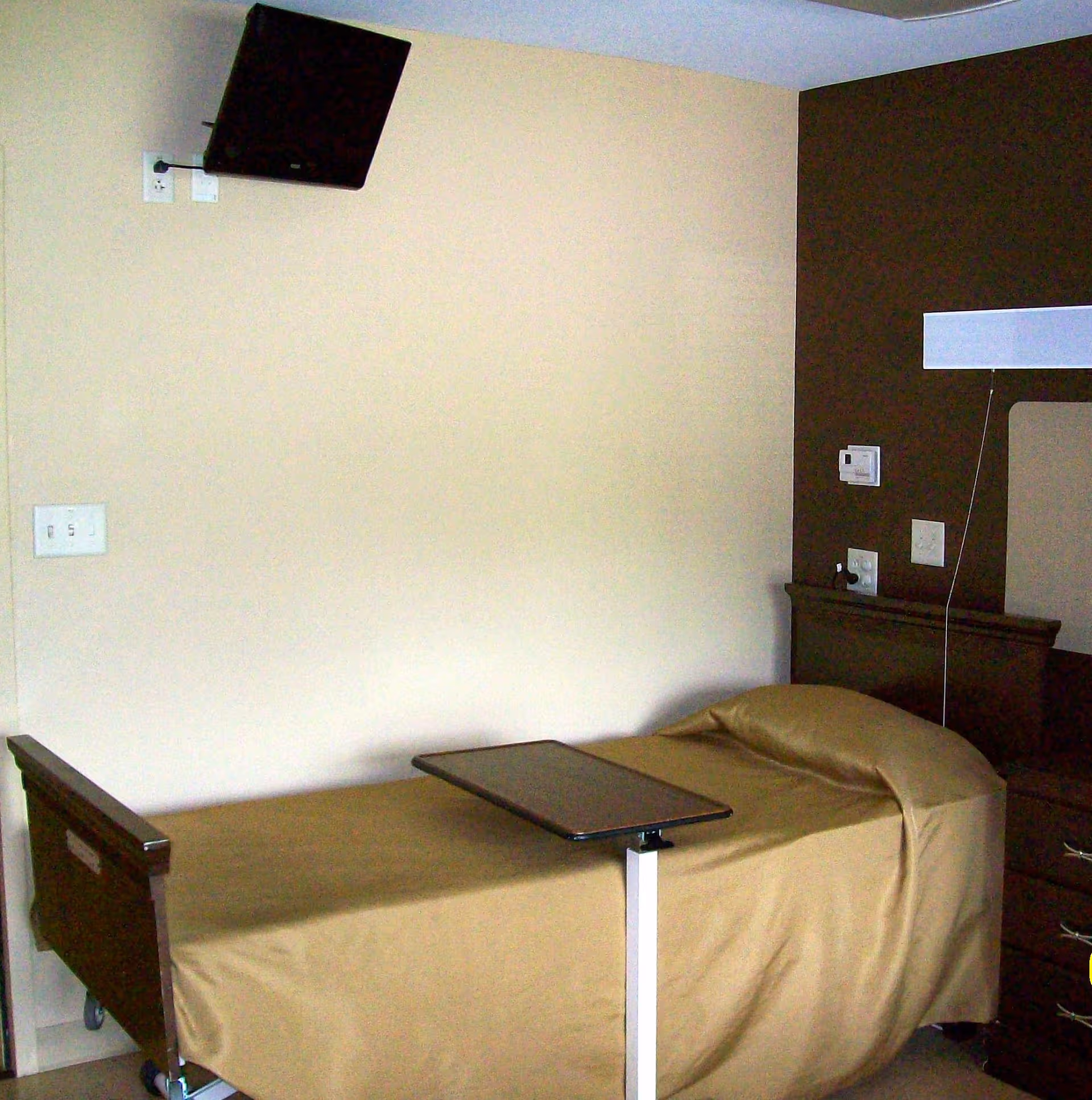A single bed with a beige bedspread in a room with a mounted flat-screen TV on the wall above the bed. There is a small adjustable overbed table positioned on the bed. The wall behind the bed is painted in two tones, beige and dark brown. A nightstand with drawers is visible next to the bed.
