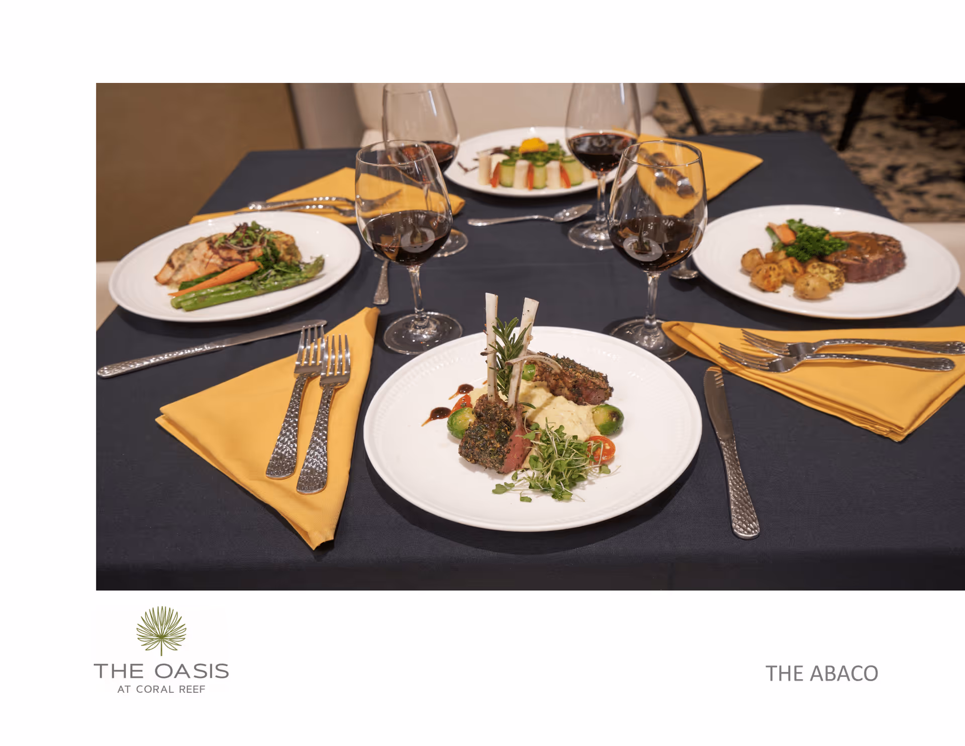 A dining table set with four plates of gourmet meals, each accompanied by a glass of red wine. The table has a dark blue tablecloth and yellow napkins with silverware neatly arranged. The meals include a variety of vegetables, meats, and garnishes.