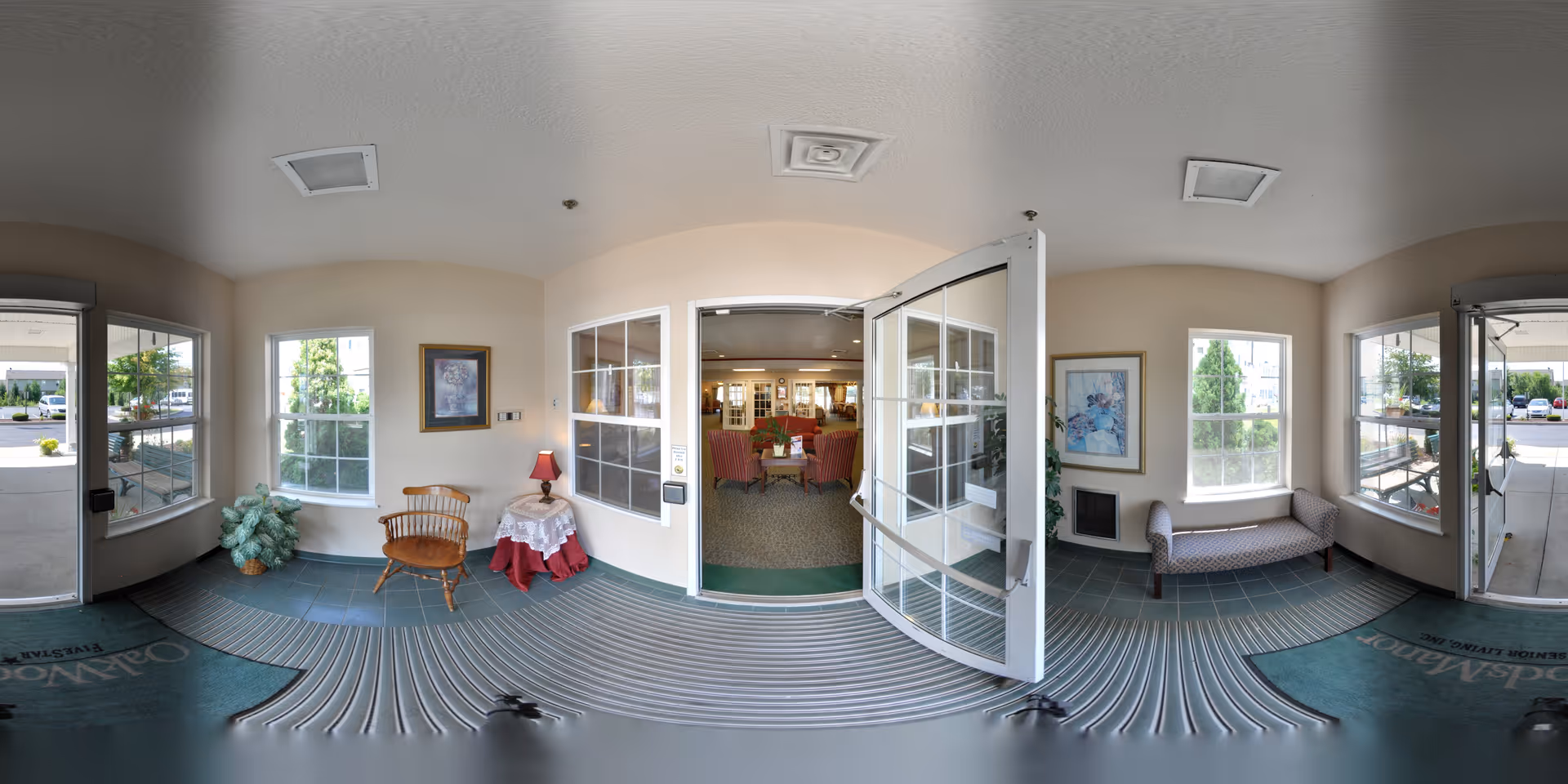 Entrance area of Oak Woods Manor showing double glass doors leading into a common area with chairs and tables. The entrance has large windows on both sides, a wooden chair with a small table and lamp, a cushioned bench, framed artwork on the walls, and welcome mats on the floor.