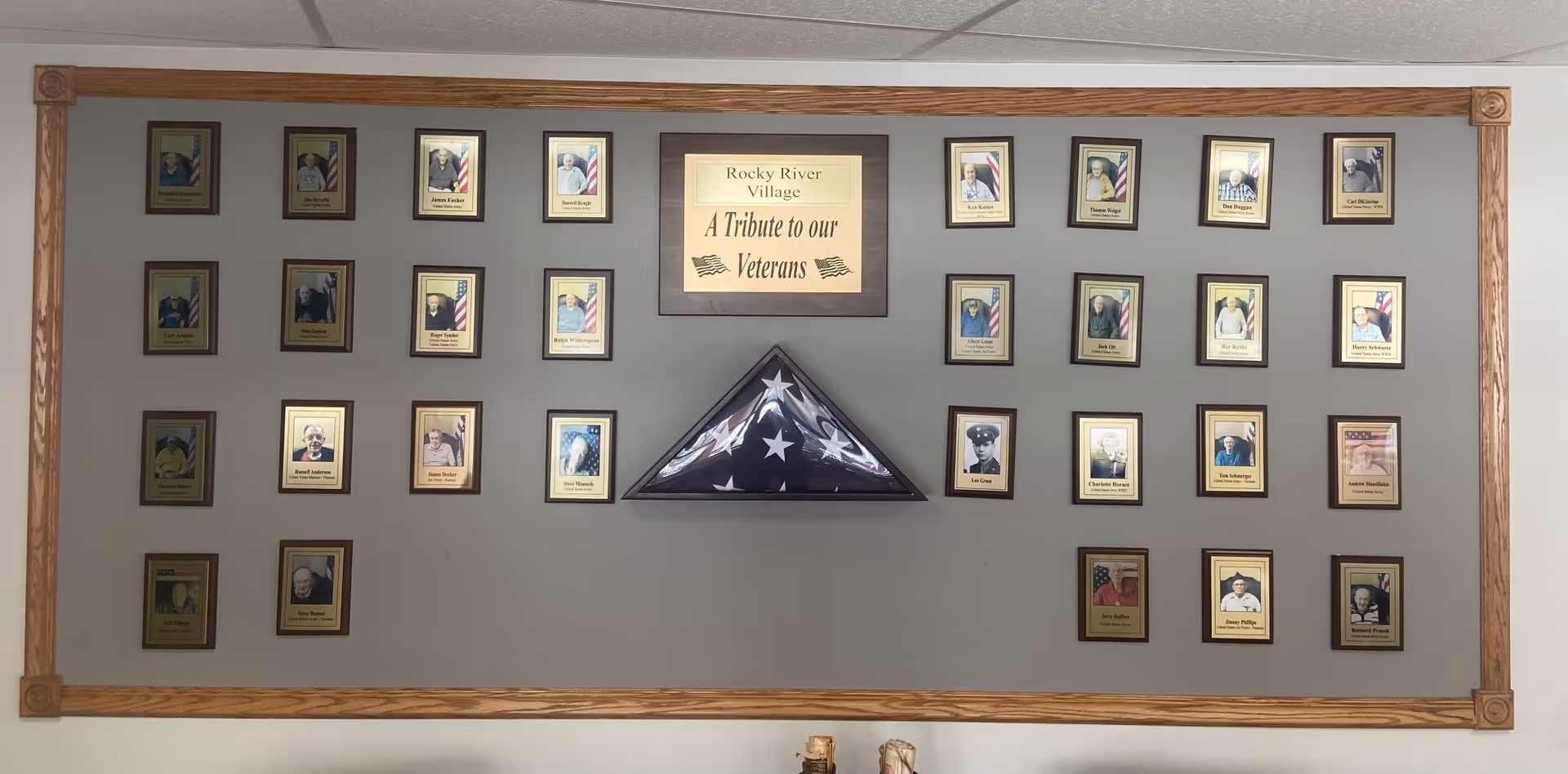 Wall-mounted memorial display of framed photos and a triangular folded American flag with a central plaque reading "A Tribute to our Veterans".