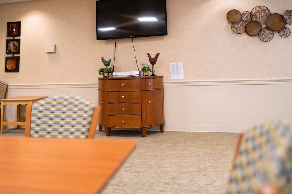 Interior view of a common area with a wooden cabinet topped with decorative items and a flat-screen TV mounted on the wall above it. The room has patterned wallpaper, carpeted floor, and partially visible chairs and tables with patterned upholstery.