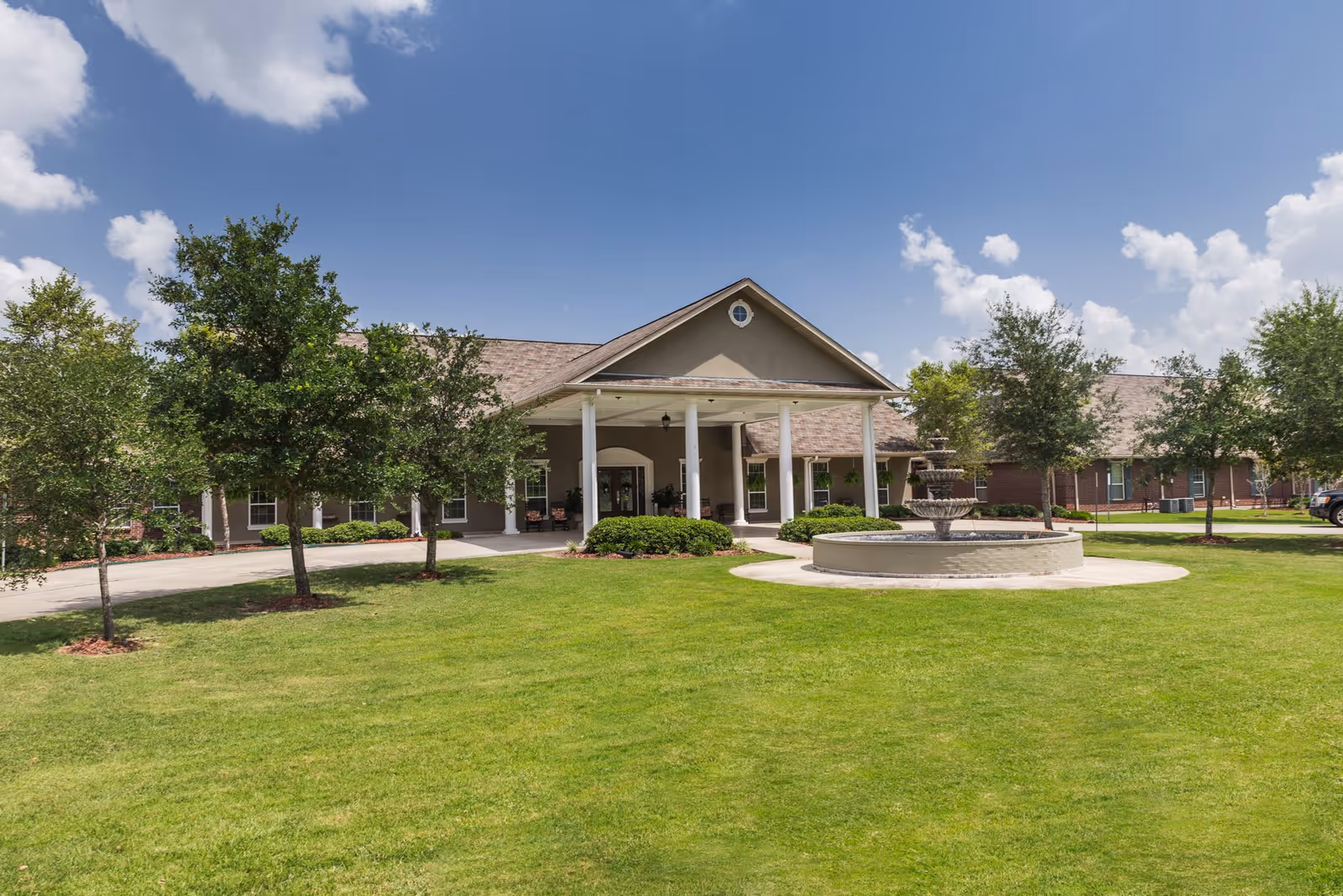 Front exterior of a single-story senior living building with a covered columned entrance, circular fountain, and manicured lawn.