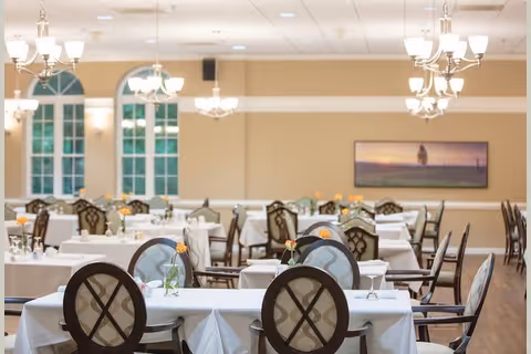 A spacious dining room with multiple tables covered in white tablecloths, each set with glassware and small flower vases. The room features elegant chandeliers hanging from the ceiling, large windows with arched tops, and a framed landscape painting on the far wall. The chairs have cushioned backs with a decorative pattern.