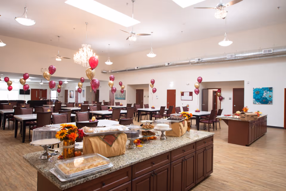 A spacious dining room decorated with maroon and gold balloons tied to the backs of chairs. The room features multiple tables and chairs arranged neatly, with two large granite countertops in the foreground displaying various covered baked goods and floral centerpieces. The ceiling has several hanging light fixtures and ceiling fans, and the floor is wood-patterned.