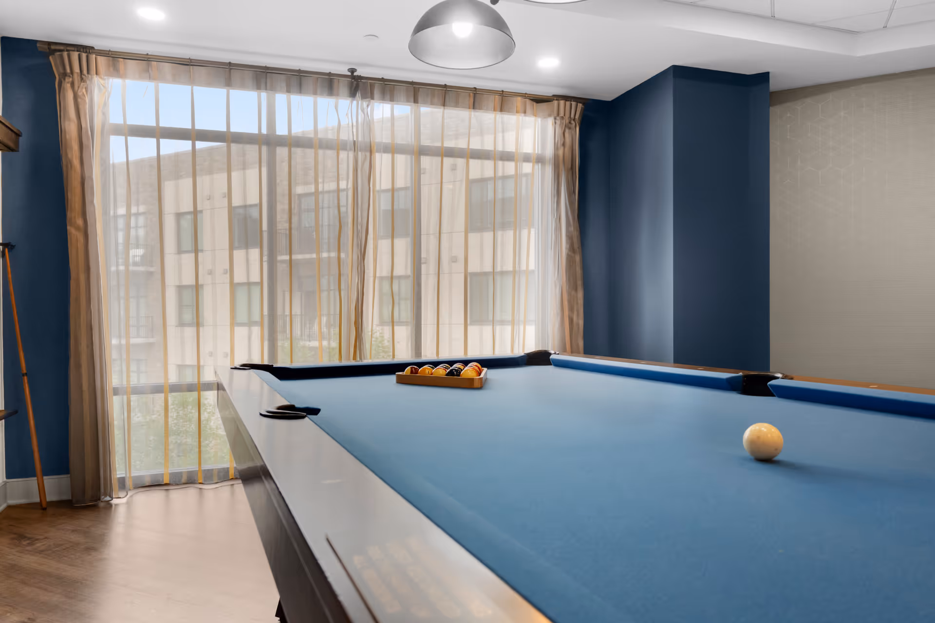 Blue-felt pool table in a bright interior game room with large windows and sheer curtains.