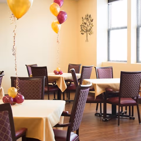 A dining area with several tables covered in beige tablecloths and surrounded by purple cushioned chairs. Each table has balloon decorations in gold and purple colors. There is a decorative tree wall art on a light-colored wall and large windows letting in natural light.