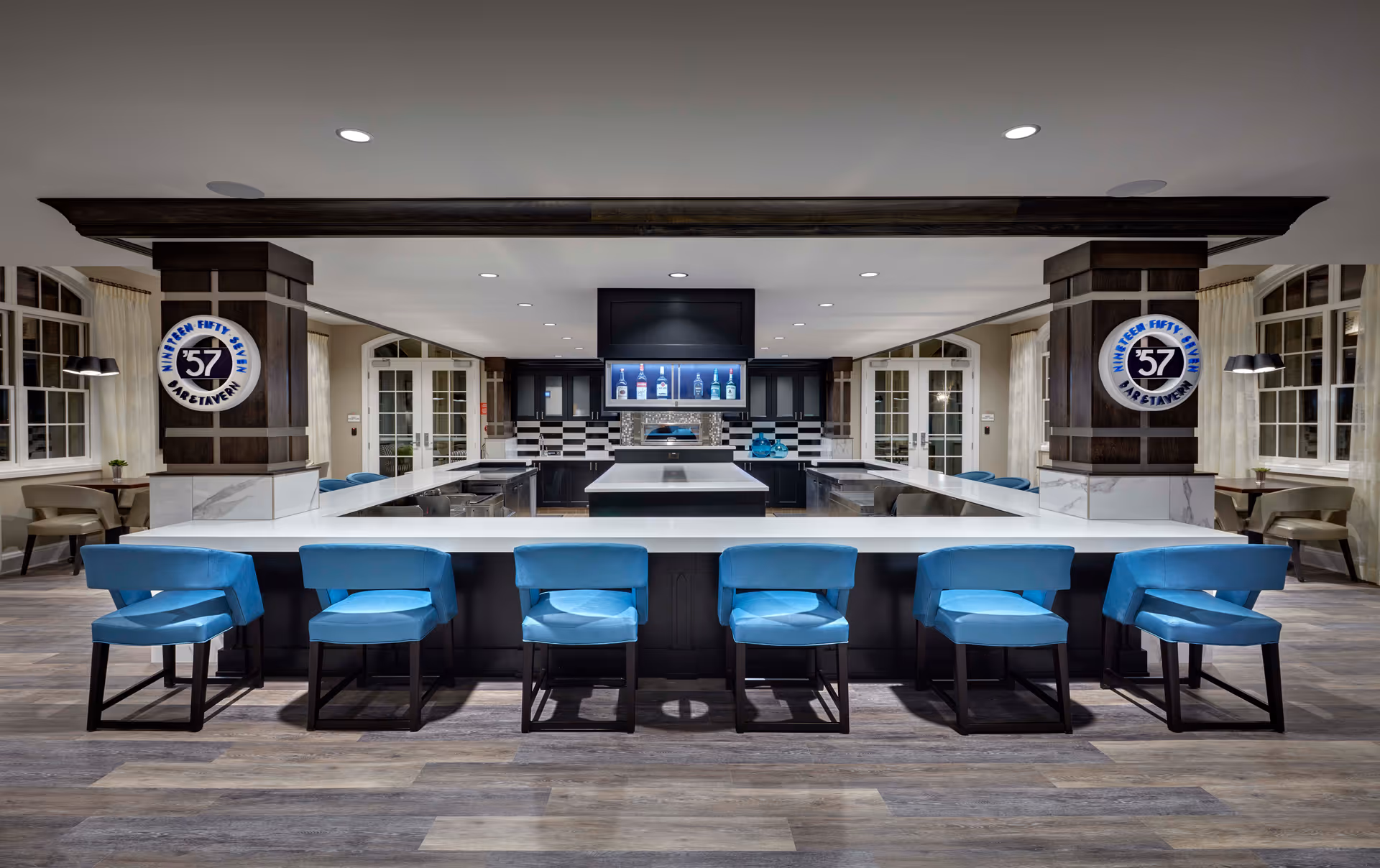 Modern bar area with a large white countertop surrounded by seven blue cushioned bar stools. The bar features dark cabinetry and a central island with a stove. Two wooden pillars with circular signs reading 'Nineteen Fifty Seven Bar & Tavern' flank the bar. The room has large windows with white curtains and additional seating areas with tables and chairs on either side.