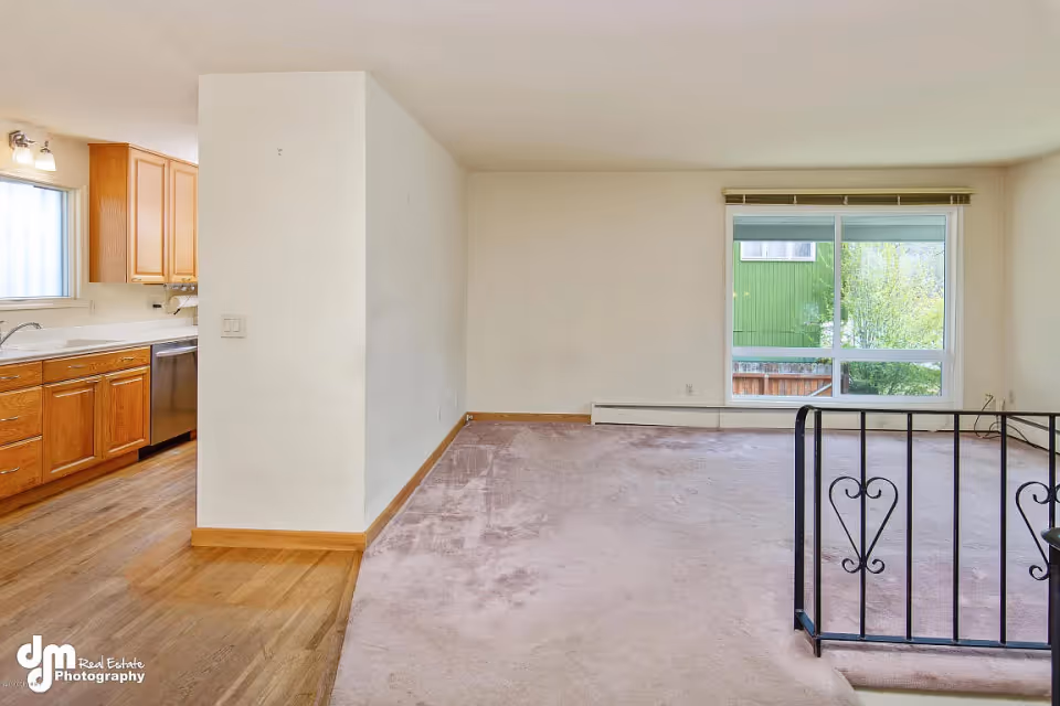 Empty living room with beige carpet, a decorative metal railing, large window, and a view into a kitchen with wooden cabinets.