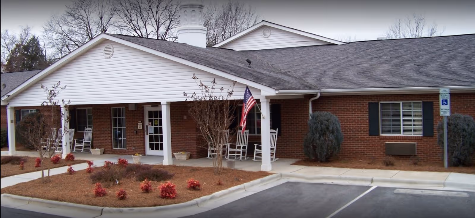 Exterior view of a single-story brick building with a white covered porch featuring rocking chairs and an American flag. There are small landscaped bushes and trees in front, a sidewalk leading to the entrance, and a handicapped parking sign visible near the building.