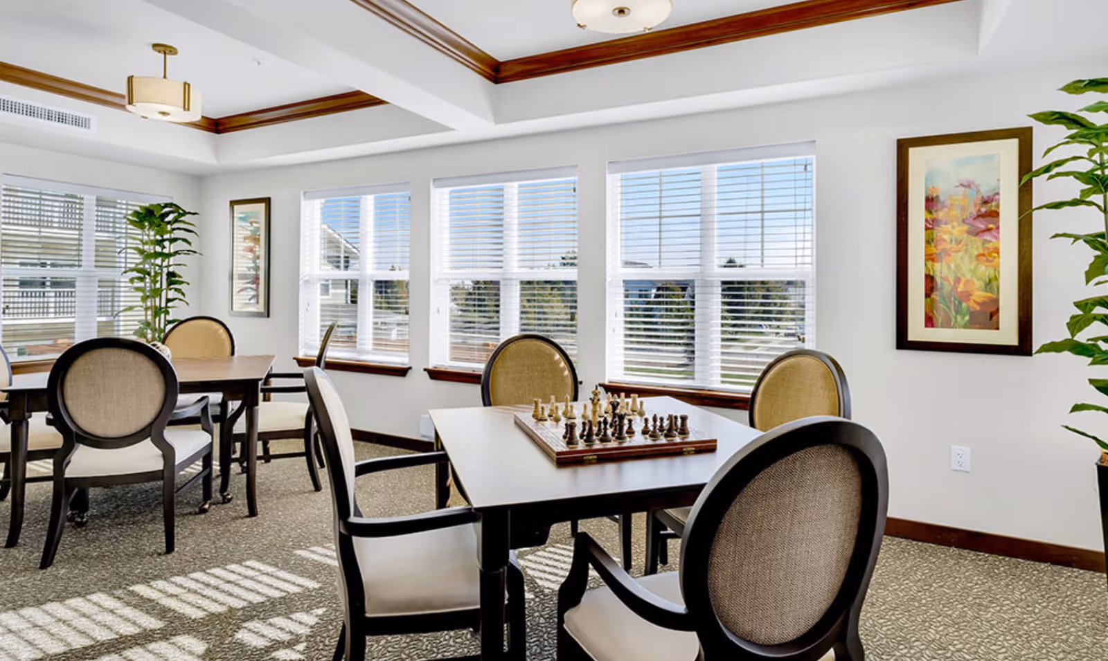 Bright communal dining and games room with tables and upholstered chairs, a chessboard on a table, large windows, plants, and framed artwork.