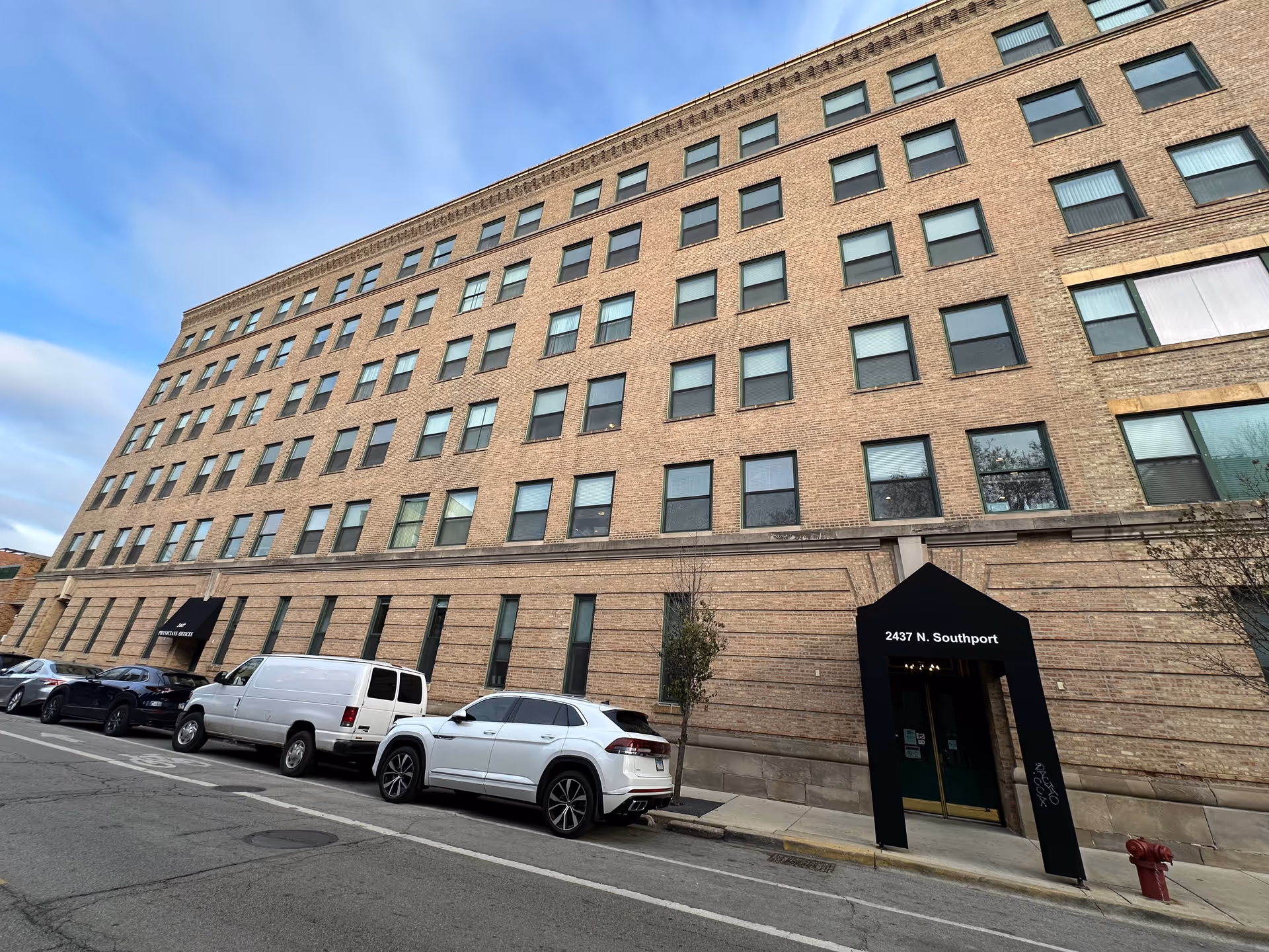 Exterior view of a multi-story brick building with many windows and two black awnings above entrances. Several cars are parked along the street in front of the building under a partly cloudy sky.