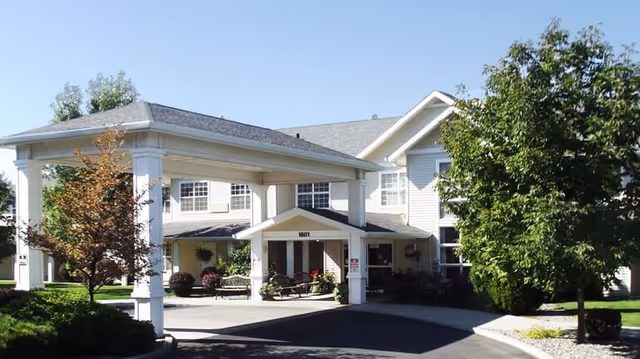 Front exterior view of McKay Creek Assisted Living facility showing a covered entrance with white pillars, a driveway, landscaped bushes and trees, and a two-story building with multiple windows.