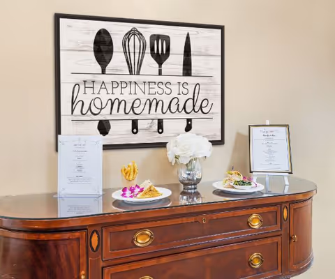 A wooden sideboard displays two plated dishes, menus and a vase of white flowers beneath a framed sign reading "Happiness is homemade."