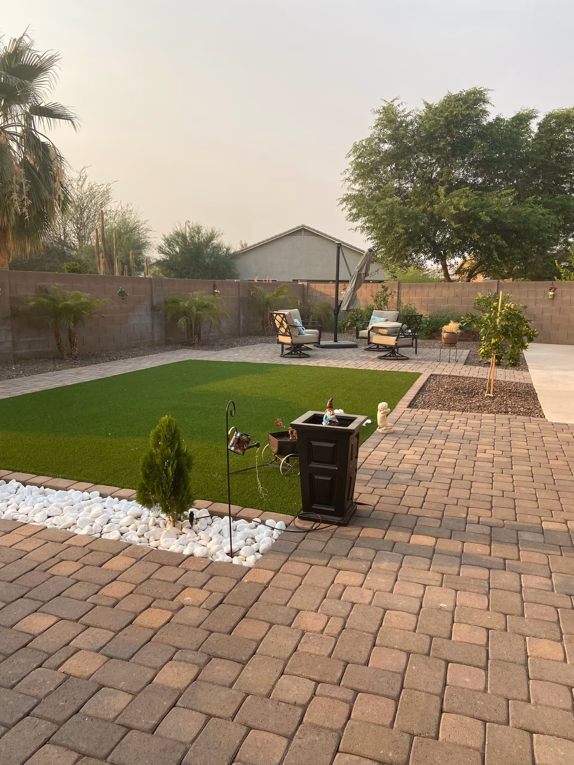 Outdoor patio area with a paved brick floor, a small patch of artificial grass bordered by white decorative stones, and several small plants. There are two cushioned chairs with a small table and an umbrella in the background, surrounded by a brick wall and trees.