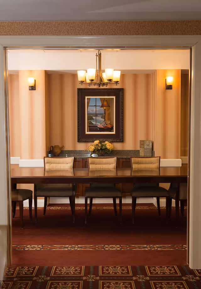 A formal dining room with a long wooden table and six cushioned chairs. The walls have vertical striped wallpaper in warm tones, and there is a framed painting of a window with a lamp and a coastal view hanging on the wall. A chandelier with six lights hangs above the table, and there are two wall sconces on either side of the painting. A floral arrangement and decorative items are placed on a sideboard against the back wall.