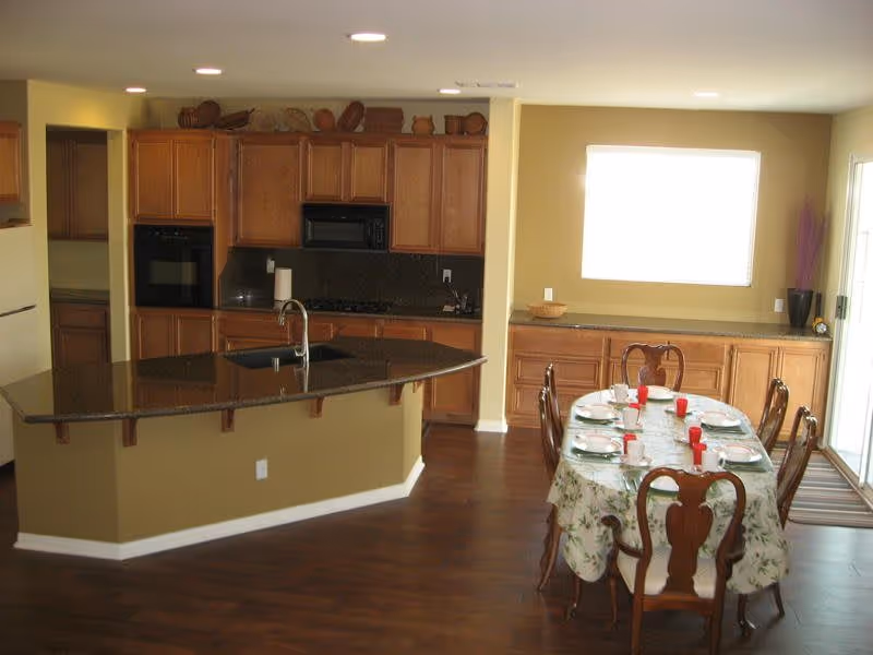 Interior view of a kitchen and dining area in a senior living facility. The kitchen features wooden cabinets, a black countertop island with a sink and faucet, a built-in oven, and a microwave. The dining area has a table covered with a floral tablecloth, set with plates, cups, and red napkins, surrounded by wooden chairs. There is a window with blinds and a sliding glass door letting in natural light.