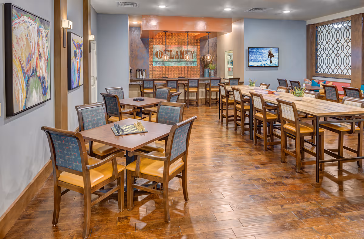A spacious dining area with wooden floors and multiple tables and chairs. There are two square tables with four chairs each in the foreground, one with a chessboard on it. In the background, there is a long table with several chairs and a bar area with stools. The walls are decorated with colorful paintings of horses and a large sign that reads 'DELANEY'. A TV screen on the wall displays an image of a person skiing. The room is well-lit with ceiling lights and wall sconces.