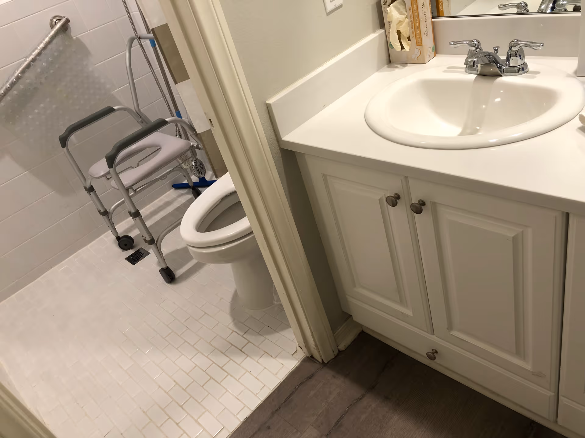 A bathroom with a white tiled floor and walls, featuring a toilet and a shower area equipped with a shower chair with wheels and a grab bar. Adjacent to the bathroom is a white sink with a countertop and cabinet below, along with a tissue box on the counter.