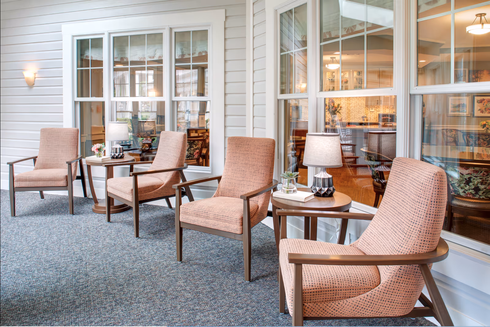 A cozy indoor seating area with four cushioned armchairs arranged around two small round wooden tables. Each table has a lamp and a small decorative item. Large windows behind the chairs reveal a warmly lit interior space with more seating and artwork on the walls.