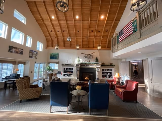 A spacious living room with a high wooden vaulted ceiling and recessed lighting. The room features a fireplace centered on the far wall, flanked by built-in shelves decorated with small trees and a model sailboat. Comfortable seating includes two blue armchairs, a red armchair, and a patterned beige armchair arranged around a coffee table on a large area rug. Large windows and glass doors allow natural light to fill the room. An American flag hangs on the upper balcony railing to the right.