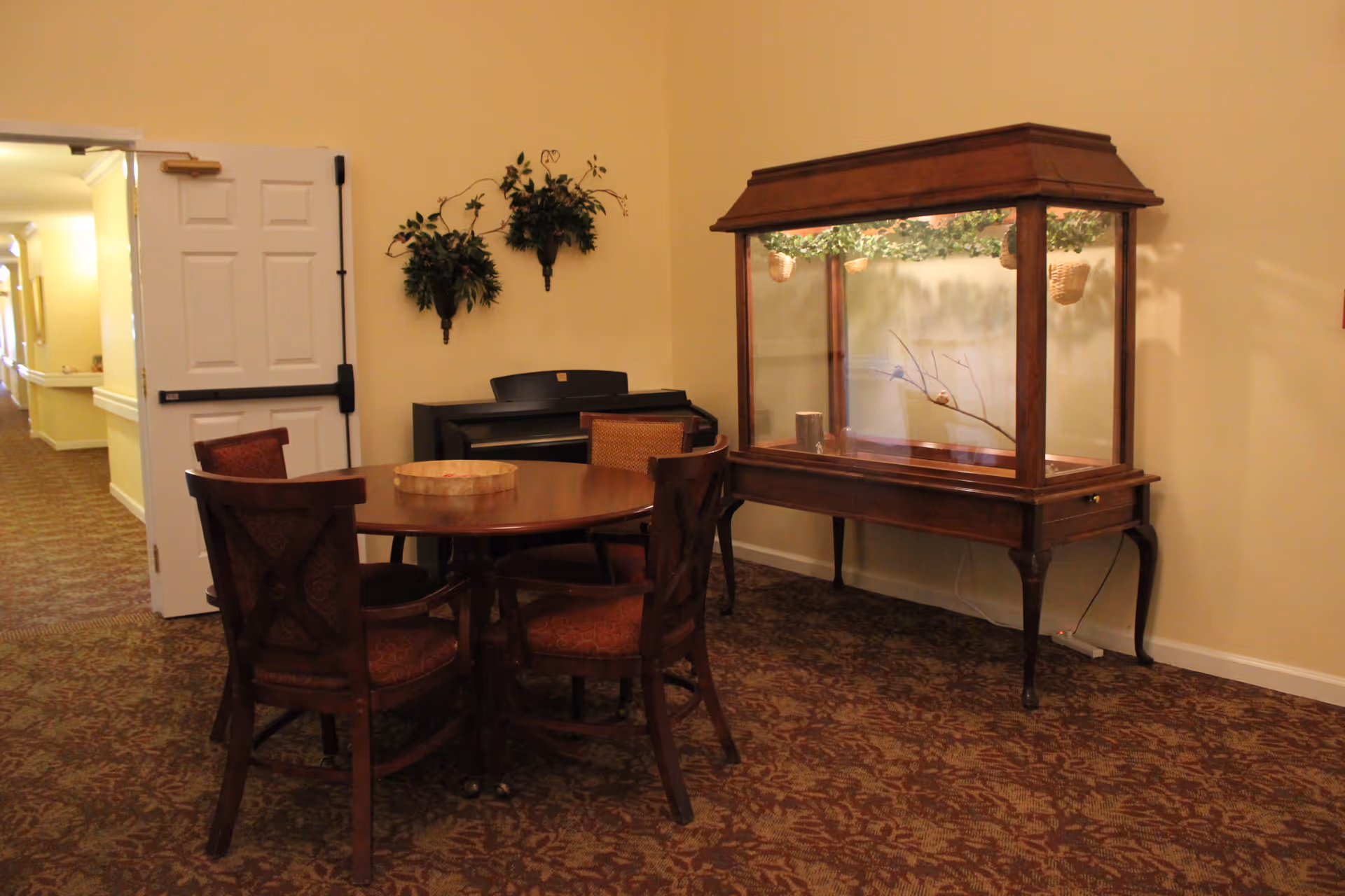 A cozy interior corner of Providence Place Assisted Living featuring a round wooden table with four upholstered chairs, a decorative wooden display case with glass panels containing branches and hanging baskets, a black piano against the wall, and two wall-mounted green plant arrangements. The area has patterned carpet flooring and cream-colored walls, with an open white door leading to a hallway.