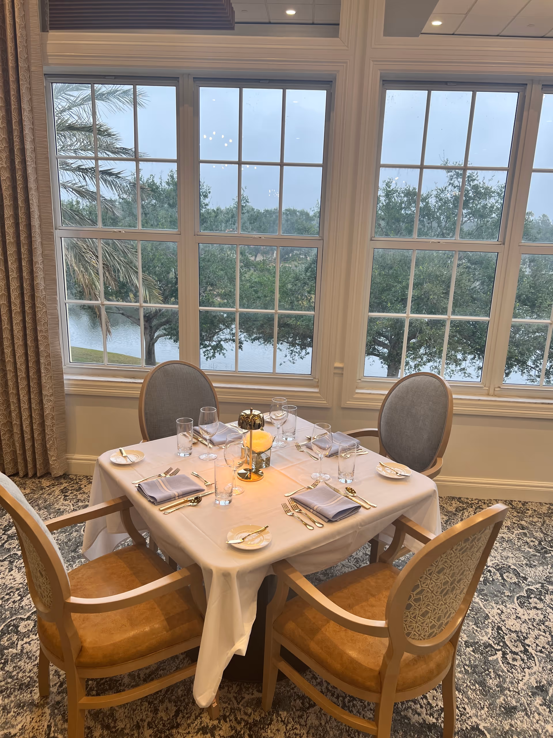 A dining table set for four with white tablecloth, napkins, glasses, plates, and silverware in front of large windows showing a view of trees and a body of water outside.