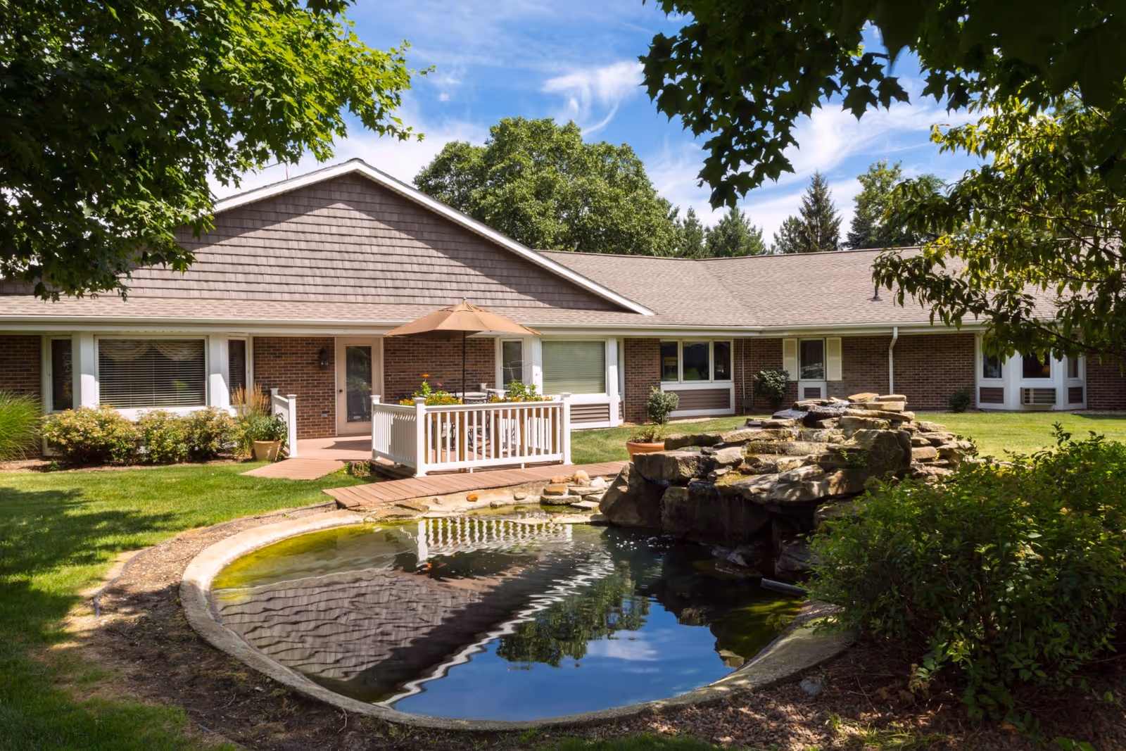 Single-story brick building with a small deck and umbrella, landscaped lawn and a pond with a rock waterfall.