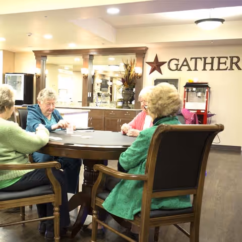 Four elderly women sitting around a round wooden table playing cards in a well-lit common area with a decorative star and the word 'GATHER' on the wall behind them. The room has wooden flooring, a counter with cabinets, and a popcorn machine in the background.