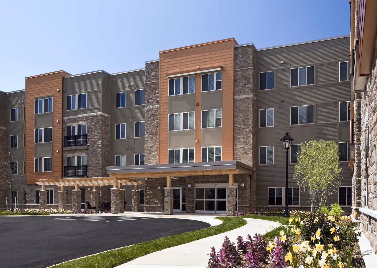 Exterior view of a modern multi-story senior living facility building with stone and wood panel accents, large windows, a covered entrance, landscaped flower beds, and a curved driveway.