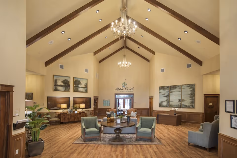 Lobby-style common room with vaulted ceilings and chandeliers, seating grouped around a central table, wood floors and wall art.