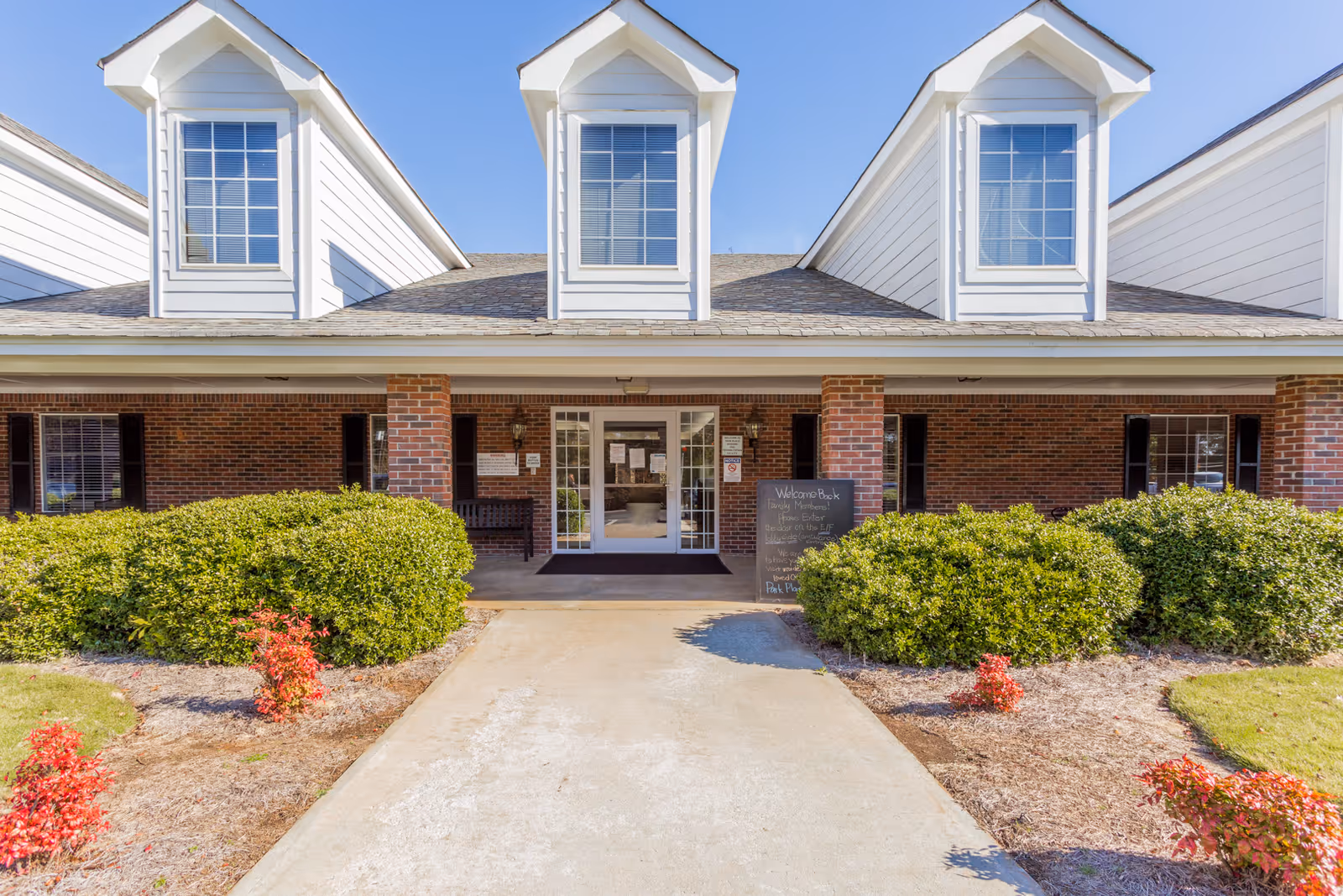 Front exterior view of Park Place Nursing and Rehab Facility showing a brick building with three dormer windows, a covered entrance with glass double doors, bushes and small red plants along the walkway leading to the entrance, and a chalkboard sign near the door.
