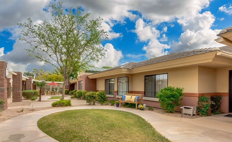 Outdoor courtyard area of a senior living facility with a curved walkway, green grass, trees, bushes, and a bench with cushions. The building has beige walls and large windows, and there is a partly cloudy blue sky above.