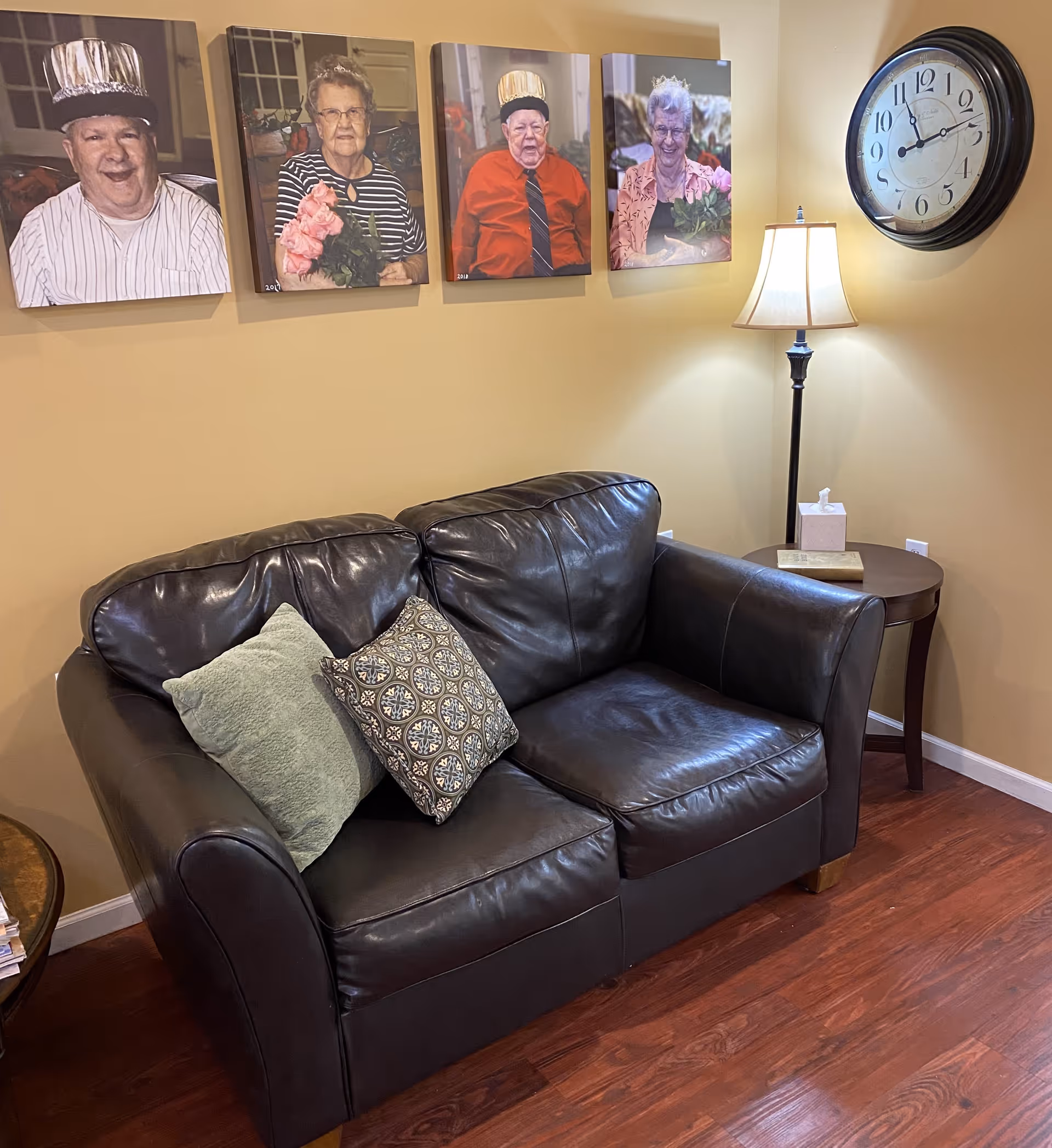 A cozy living room corner with a dark brown leather loveseat adorned with two decorative pillows. Above the loveseat are four framed photos of elderly individuals wearing festive hats and holding flowers. To the right, there is a round wooden side table with a tissue box and a book, next to a tall floor lamp with a white lampshade. A large round wall clock hangs on the beige wall above the lamp.