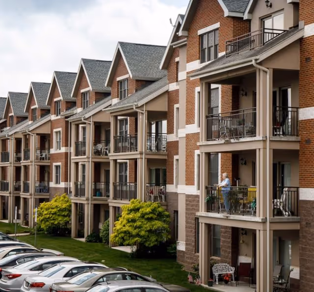 Brick multi-story senior living building with rows of balconies, gabled rooflines, and parked cars in front.
