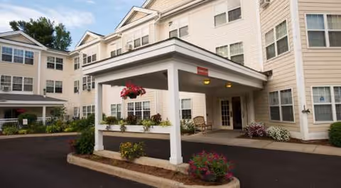 Two-story beige senior living building with a covered entrance and colorful flower planters.